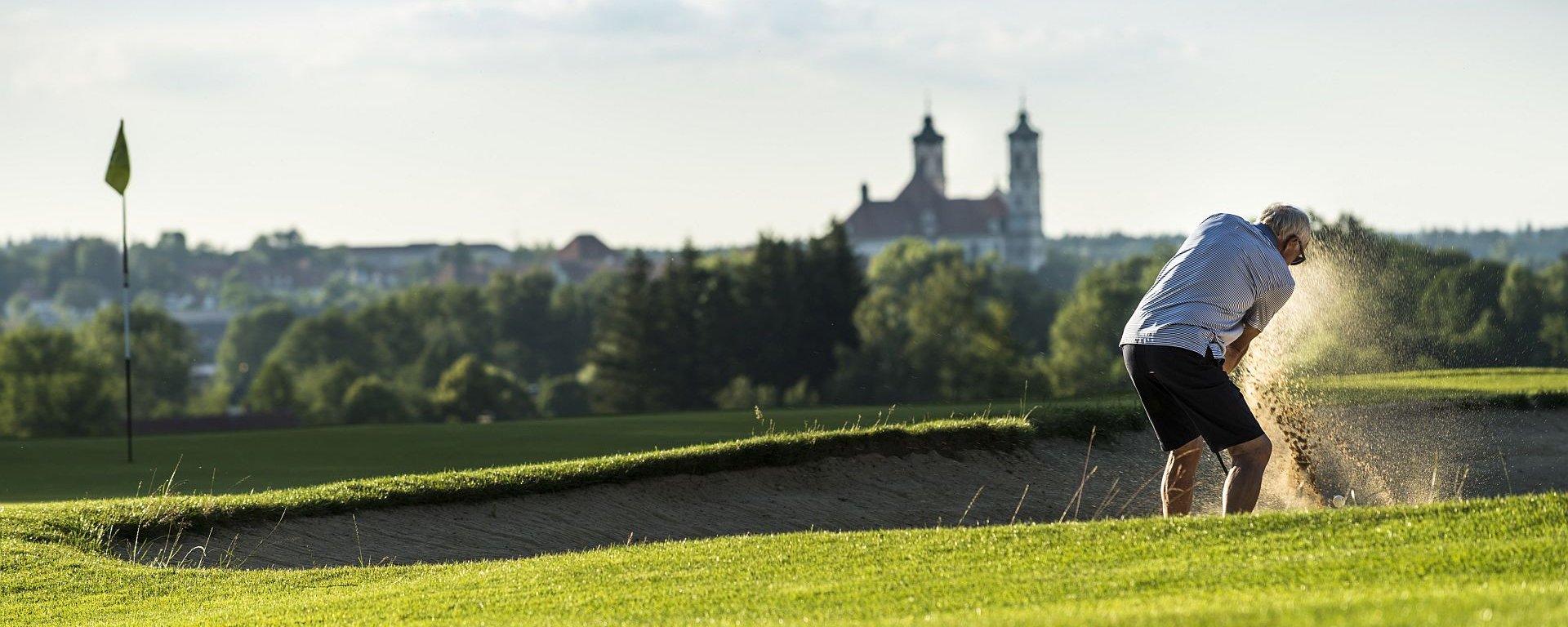 Ein Golfer führt einen Bunkerschlag in einem Sandbunker nahe des Lochs auf dem Golfplatz des Allgäuer Golf- und Landclub e.V. in Ottobeuren aus. Im Gegenlicht der untergehenden Sonne fliegen in Ballnähe Sandkörner durch die Luft (Credit: Stefan von Stengel)