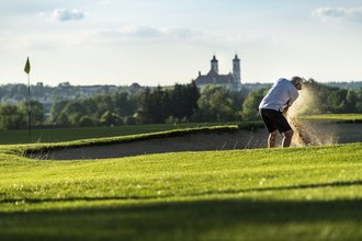 Ein Golfer führt einen Bunkerschlag in einem Sandbunker nahe des Lochs auf dem Golfplatz des Allgäuer Golf- und Landclub e.V. in Ottobeuren aus. Im Gegenlicht der untergehenden Sonne fliegen in Ballnähe Sandkörner durch die Luft (Credit: Stefan von Stengel)