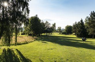 Panoramaaufnahme eines Kurses des Golfanlage des Allgäuer Golf- und Landclub e.V.: Ein Golferpaar läuft über das gepflegte Green  – tief im Hintergrund ragt die Basilika Ottobeuren hinter den Bäumen hervor (Credit: Stefan von Stengel)