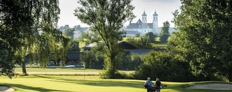 Ein Golferpaar läuft über das gepflegte Green des Golfanlage des Allgäuer Golf- und Landclub e.V. – im Hintergrund ragt die Basilika Ottobeuren hinter den Bäumen hervor (Credit: Stefan von Stengel)
