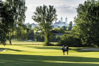 Ein Golferpaar läuft über das gepflegte Green des Golfanlage des Allgäuer Golf- und Landclub e.V. – im Hintergrund ragt die Basilika Ottobeuren hinter den Bäumen hervor (Credit: Stefan von Stengel)