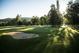 Ein Loch mit Bunker, auf dem Golfplatz des Allgäuer Golf- und Landclub e.V. – zwischen der großen Anzahl umliegender Bäume bricht das Licht der Abendsonne durch und taucht das Green in ein malerisches goldenes Licht (Credit: Stefan von Stengel)