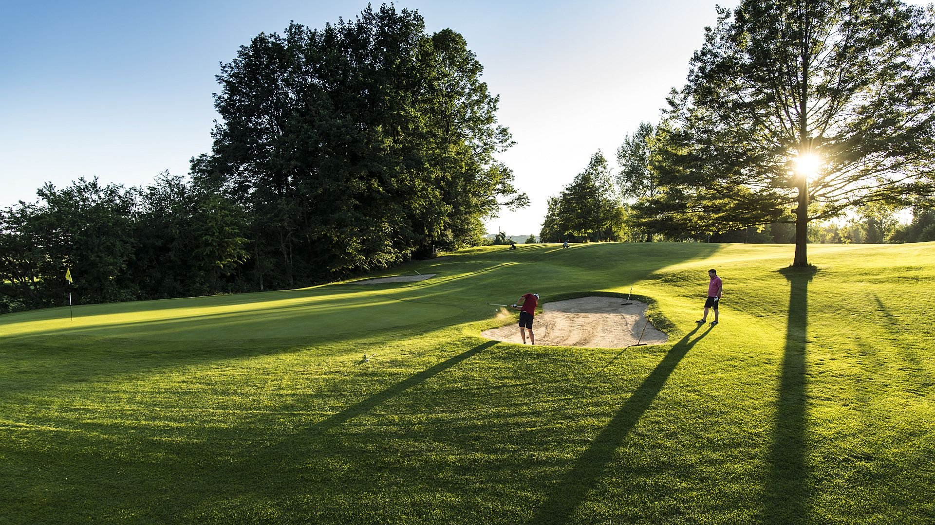 Zwei Golfer auf dem Golfplatz des Allgäuer Golf- und Landclub e.V., von denen einer einen Bunkerschlag in einem Sandbunker nahe des Lochs ausführt. (Credit: Stefan von Stengel)