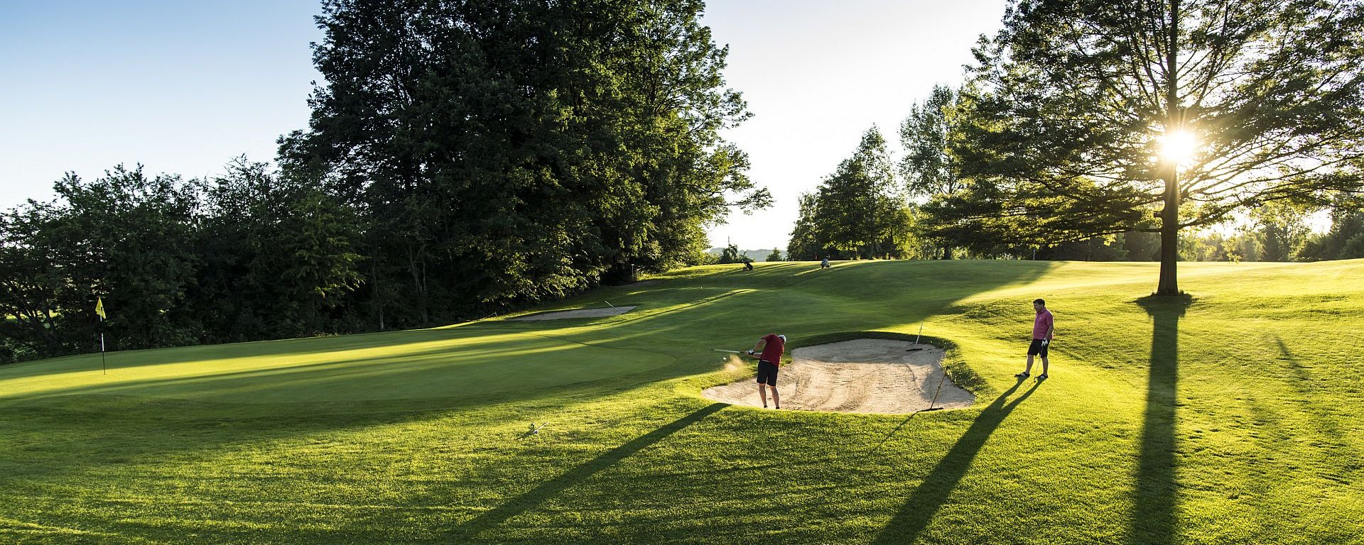 Zwei Golfer auf dem Golfplatz des Allgäuer Golf- und Landclub e.V., von denen einer einen Bunkerschlag in einem Sandbunker nahe des Lochs ausführt. (Credit: Stefan von Stengel)