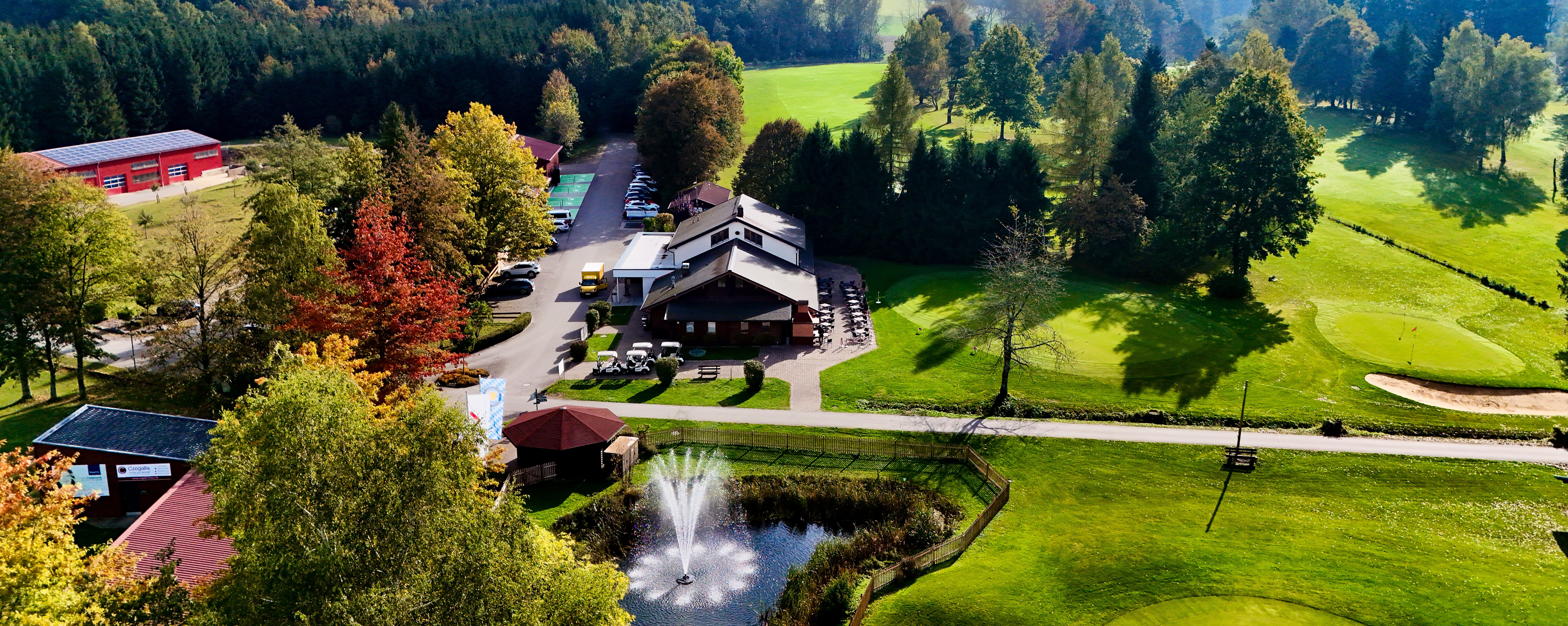 Eine Luftaufnahme des Golfplatzes. Auf der linken Seite befindet sich das Gemeinschaftshaus und Restaurant des Clubs. Davor befinden sich einige Parkplätze. Rechts sind erste Golfbahnen zu sehen und im Vordergrund befindet sich ein  Teich, der von Bäumen umringt ist.