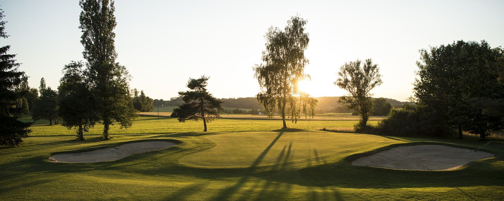 Ein Loch, flankiert von zwei Bunkern, auf dem Golfplatz des Allgäuer Golf- und Landclub e.V. – im Licht der Abendsonne werfen die Bäume sanfte Schatten auf das Green (Credit: Stefan von Stengel)