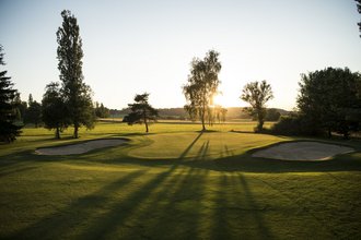 Ein Loch, flankiert von zwei Bunkern, auf dem Golfplatz des Allgäuer Golf- und Landclub e.V. – im Licht der Abendsonne werfen die Bäume sanfte Schatten auf das Green (Credit: Stefan von Stengel)