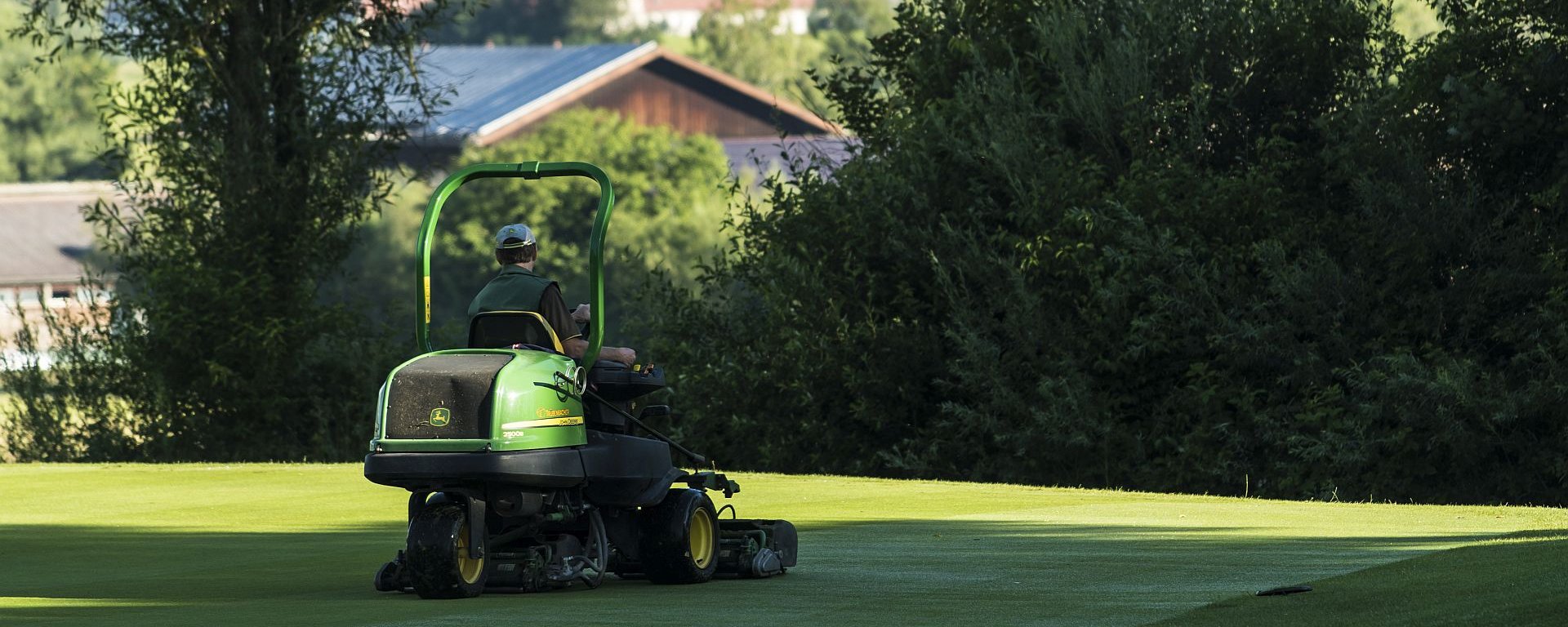 Ein Greenkeeper mäht auf seinem Mähtraktor das Green im Allgäuer Golf- und Landclub e.V. – im Hintergrund ragt die Basilika Ottobeuren hinter den Büschen hervor (Credit: Stefan von Stengel)