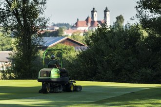 Ein Greenkeeper mäht auf seinem Mähtraktor das Green im Allgäuer Golf- und Landclub e.V. – im Hintergrund ragt die Basilika Ottobeuren hinter den Büschen hervor (Credit: Stefan von Stengel)