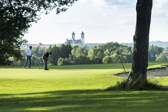 Zwei Golfer putten auf dem Green des Allgäuer Golf- und Landclub e.V. in Ottobeuren vor dem Hintergrund der hinter den Bäumen hervor ragenden Basilika (Credit: Stefan von Stengel)