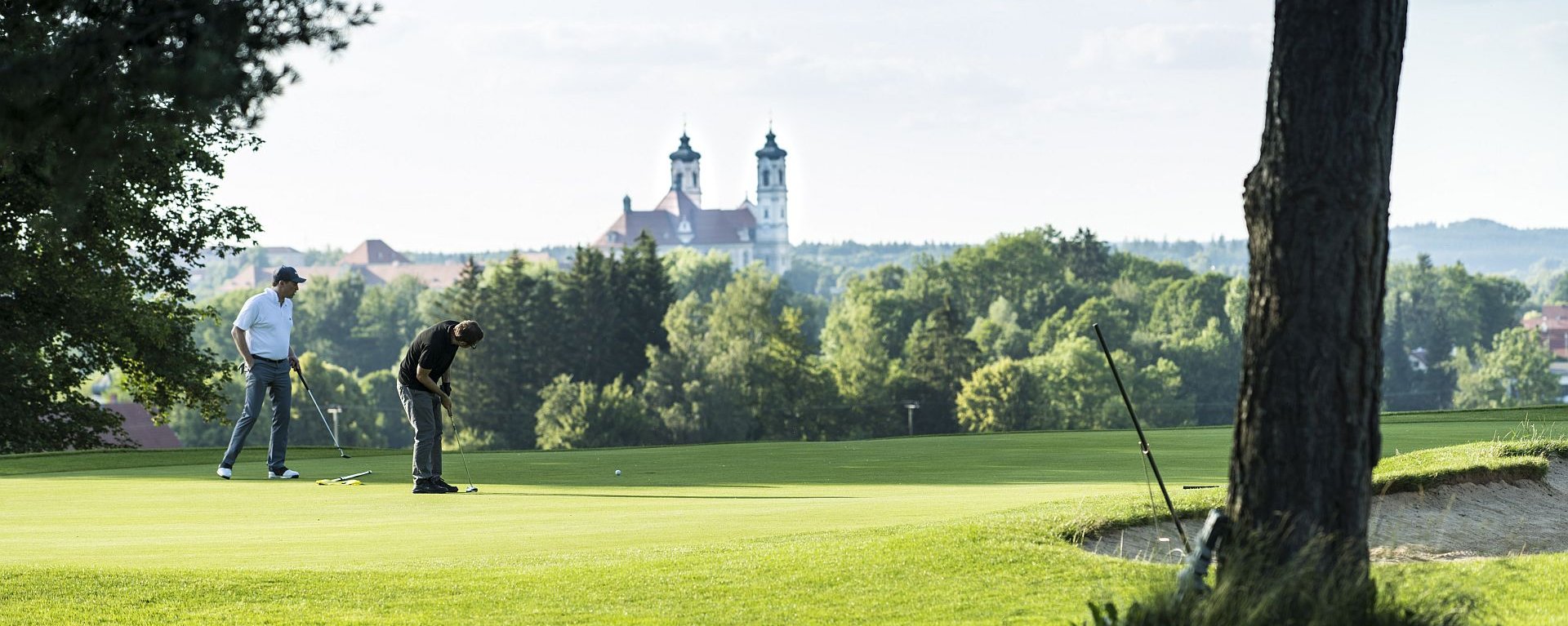 Zwei Golfer putten auf dem Green des Allgäuer Golf- und Landclub e.V. in Ottobeuren vor dem Hintergrund der hinter den Bäumen hervor ragenden Basilika (Credit: Stefan von Stengel)