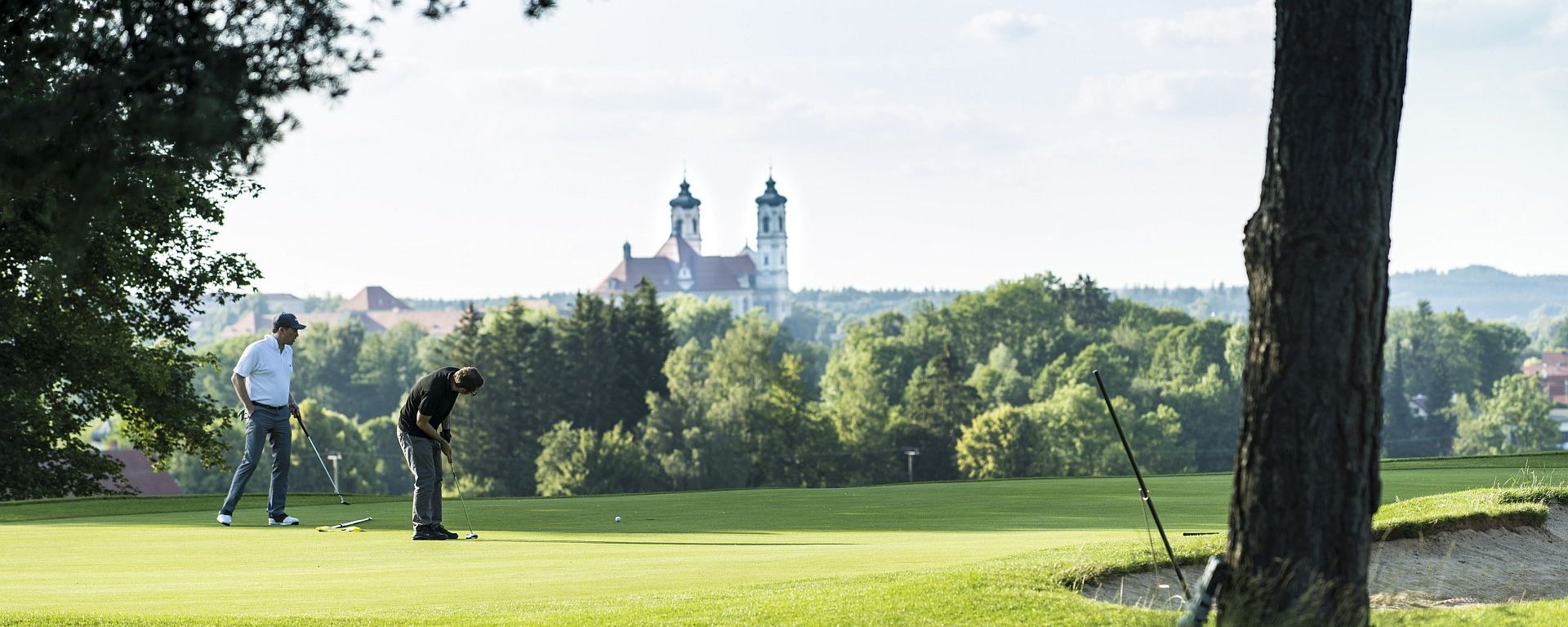 Zwei Golfer putten auf dem Green des Allgäuer Golf- und Landclub e.V. in Ottobeuren vor dem Hintergrund der hinter den Bäumen hervor ragenden Basilika (Credit: Stefan von Stengel)